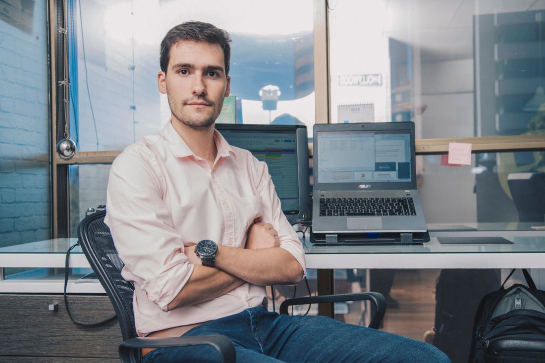 A man sits with arms crossed at an office desk, looking directly at the camera with laptop screens visible behind him.