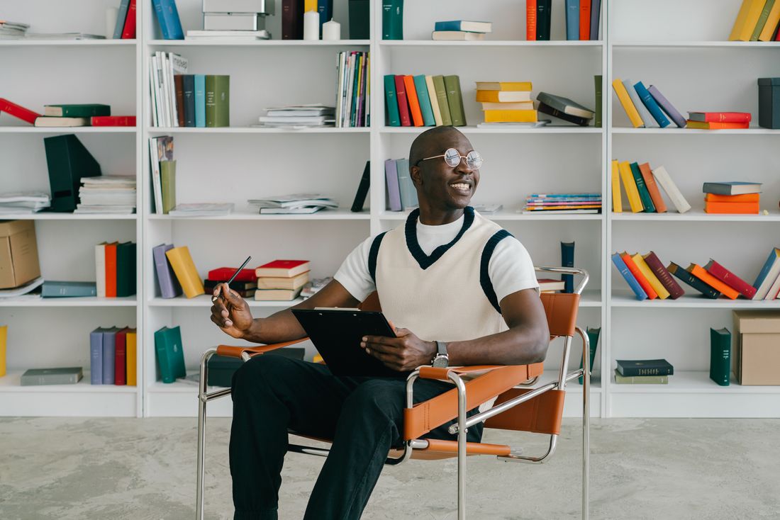 A man sits in a chair in front of a full bookshelf, smiling and mid-response as if answering a question during a relaxed conversation.