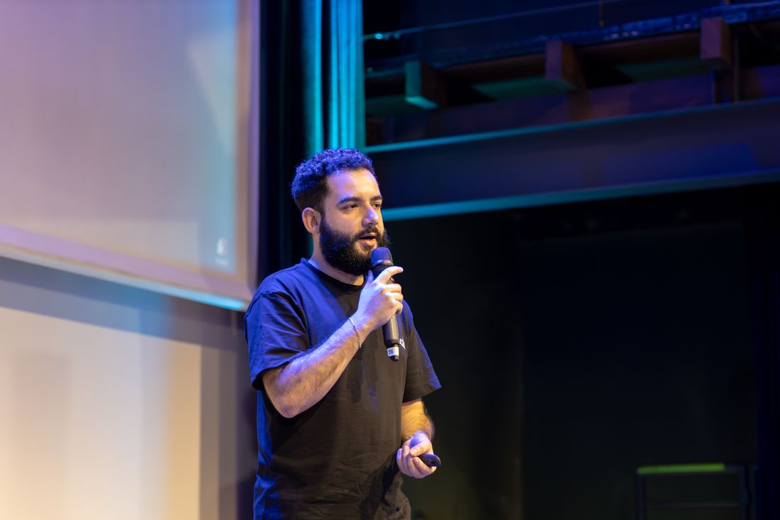 A bearded man speaks into a handheld microphone under blue stage lighting, gesturing as he talks, with a blank projection screen and dark stage rigging visible behind him.