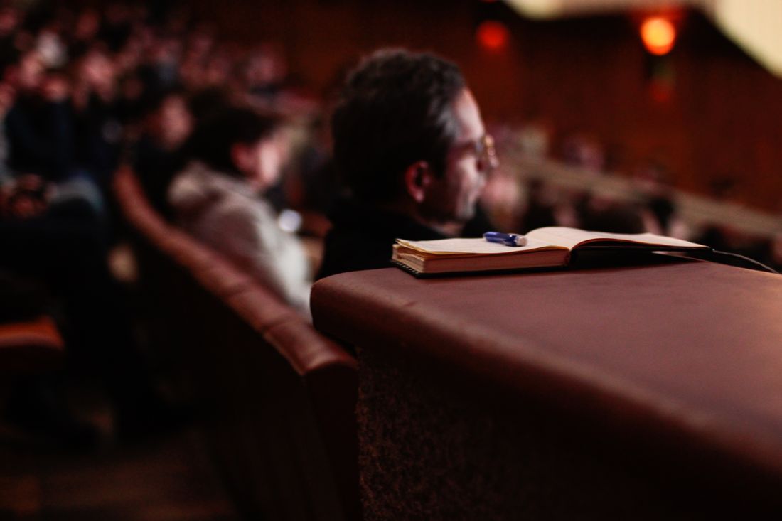 An open notebook and pen rest on the back of a theater seat in a dim auditorium, with a person seated nearby in profile and a blurred audience filling the rows behind.