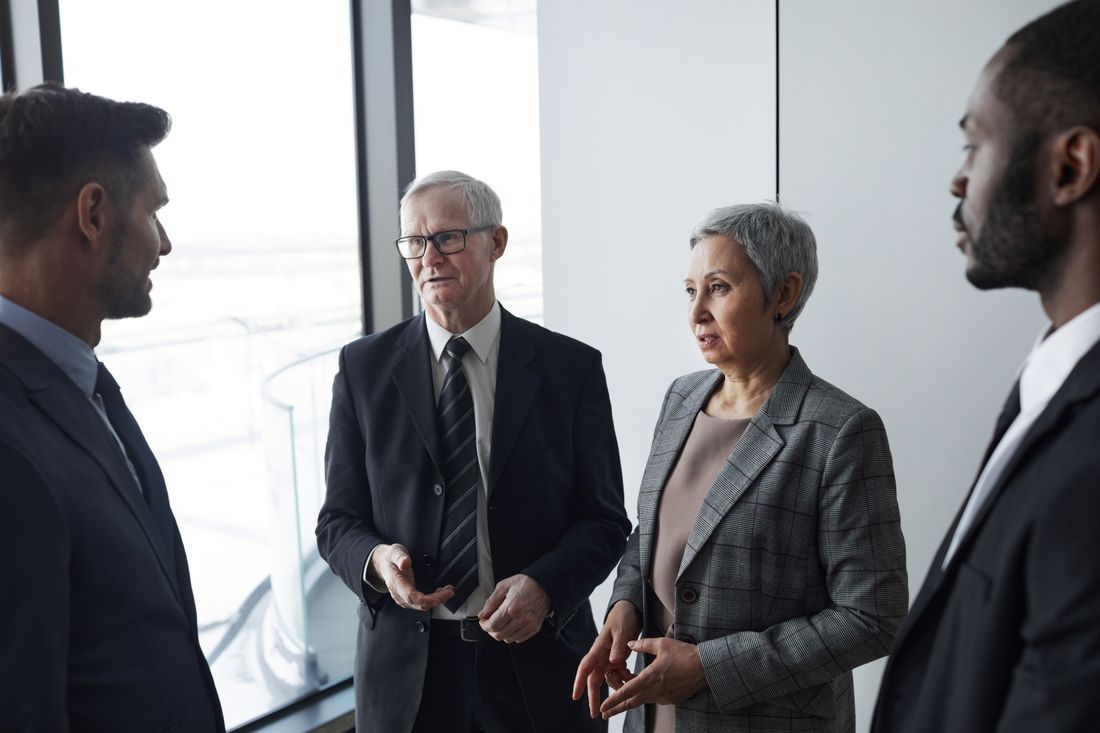 Four business professionals of different ages, in suits and a blazer, standing and talking together in a bright office hallway near a window.