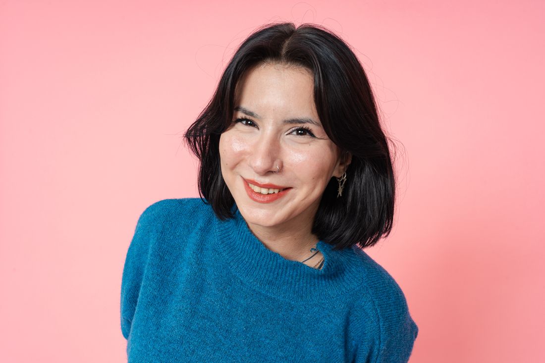 A woman with a nose ring and short dark hair, wearing a blue sweater, smiling directly at the camera against a solid pink studio backdrop.