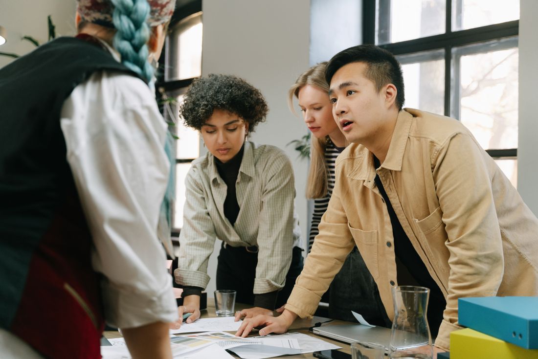 Five casually dressed colleagues leaning in together over a table covered in sketches and color swatches in a bright, window-lined office.