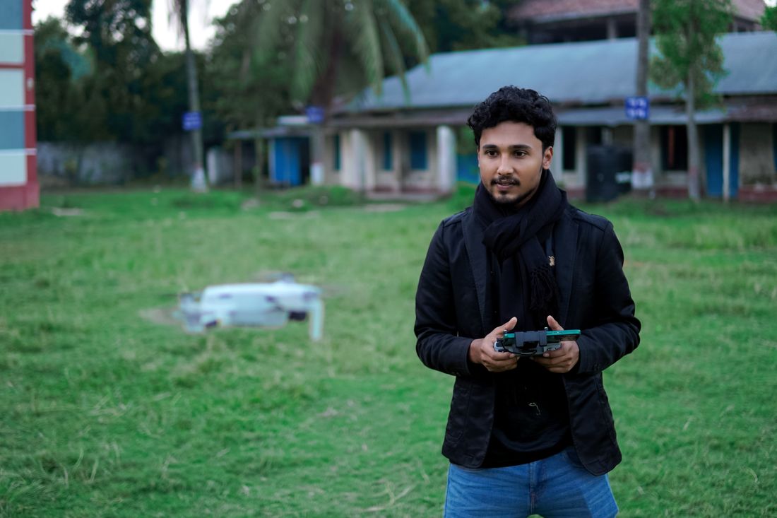 A man stands outdoors holding a drone remote controller with both hands while a small quadcopter drone hovers a few feet away on the grass, with a building softly out of focus behind him.