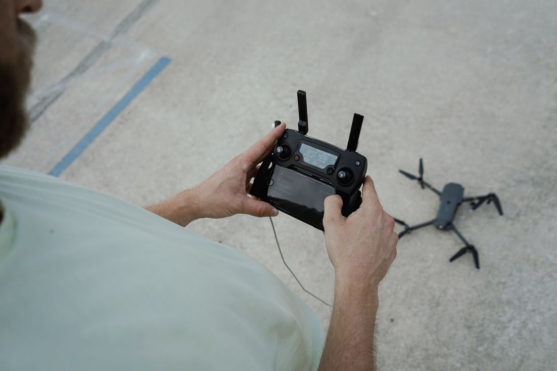 A person crouches over a drone resting on a paved outdoor surface, holding its remote controller with both hands as the drone's propellers and legs rest on the pavement nearby.