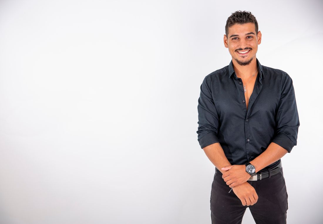 A man in a dark button-down shirt stands smiling against a plain white studio background, hands loosely clasped in front of him.