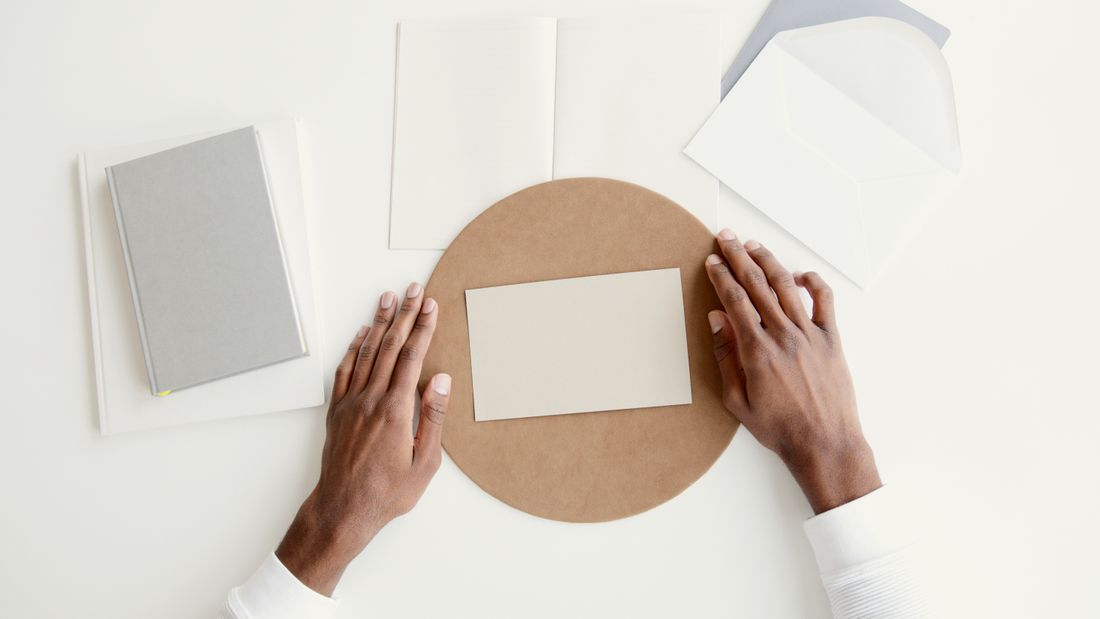 Two hands rest on either side of a blank cream card set on a round tan mat, with a blank folded card, a plain gray notebook, and an open blank envelope arranged nearby on a white table.