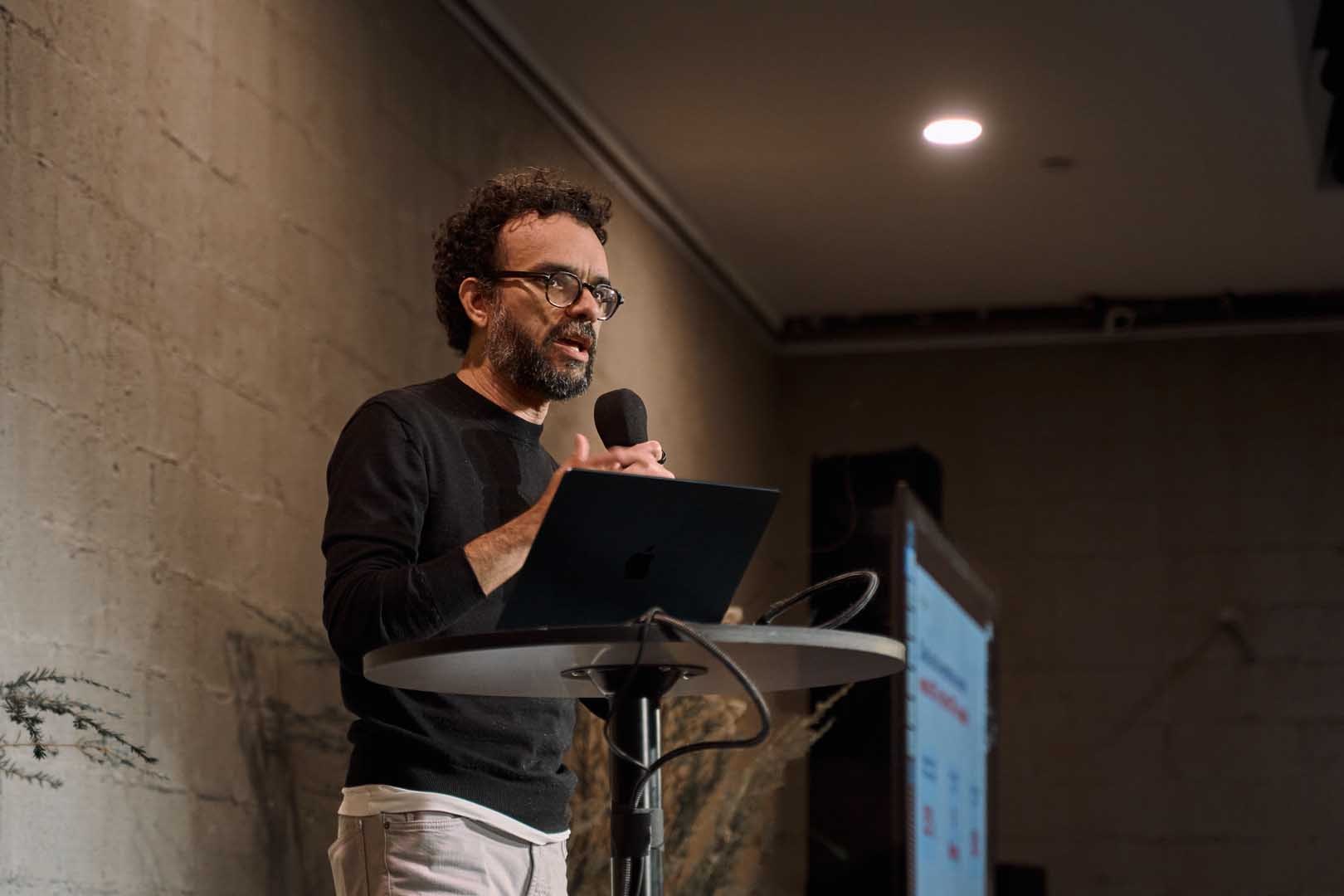 Bearded man in black sweater speaking at lectern with laptop and data screen.