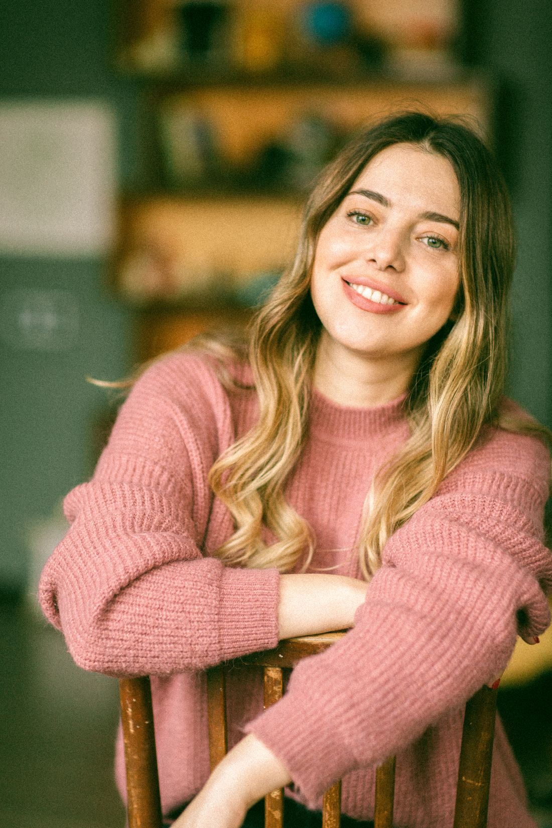 A woman in a pink sweater sitting backward on a chair, smiling warmly in natural light.