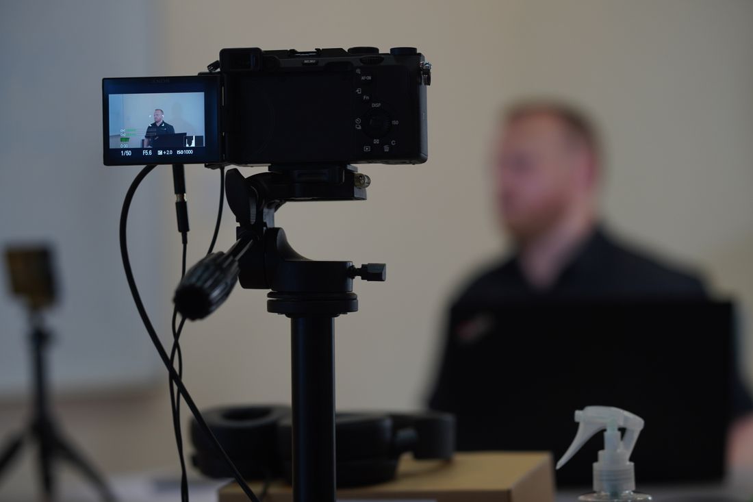 A video camera mounted on a stabilizer rig filming indoors, with a speaker blurred in the background of the room.