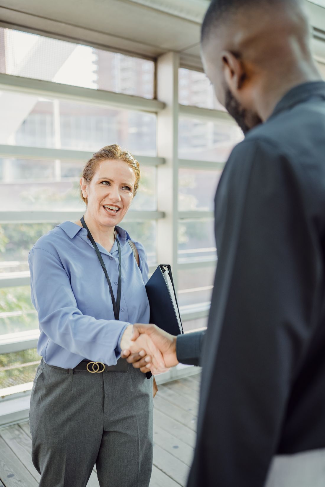 A man and a woman in business attire shaking hands in a bright office hallway.