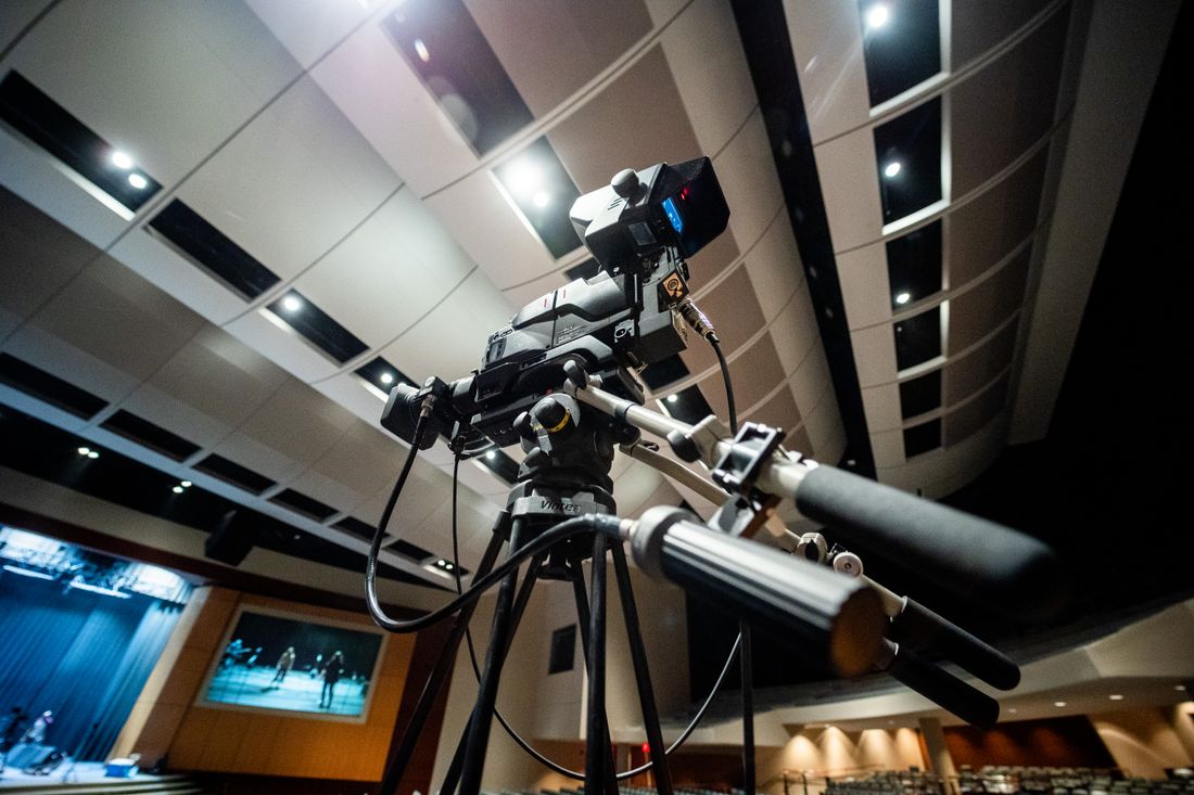 A professional video camera on a tripod set up in an empty auditorium under stage lighting, representing a crew ready ahead of an event