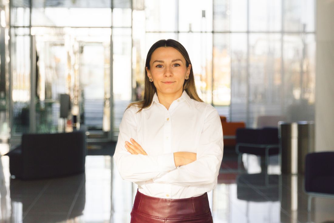 A woman stands with arms crossed in a bright, glass-walled office lobby, daylight filling the space behind her.