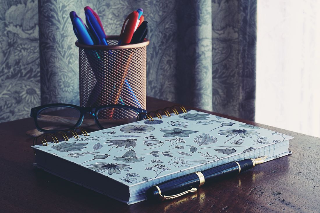 A closed floral-patterned notebook rests beside a cup of pens and reading glasses on a wooden desk near a window.