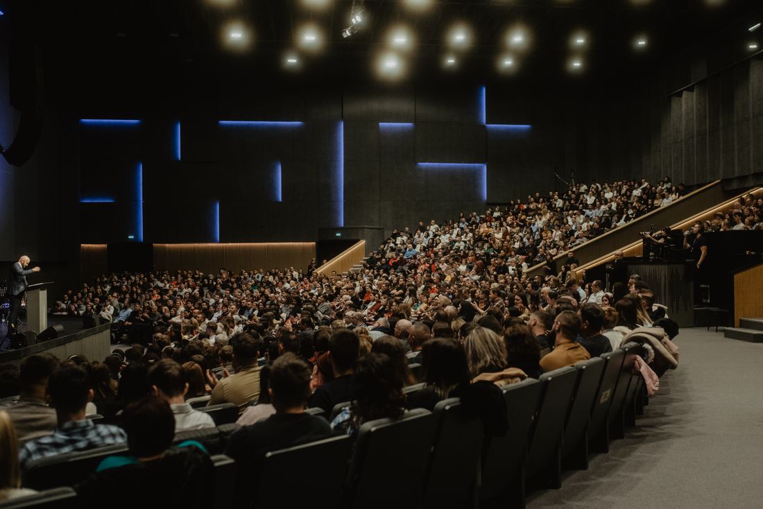 A wide, dimly lit auditorium filled with a large seated crowd, with open ceiling and wall space above the stage.