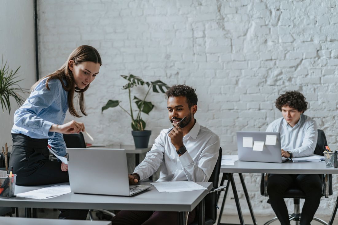 Coworkers at laptops in an open office with an exposed brick wall, one on a phone call while a colleague looks on.