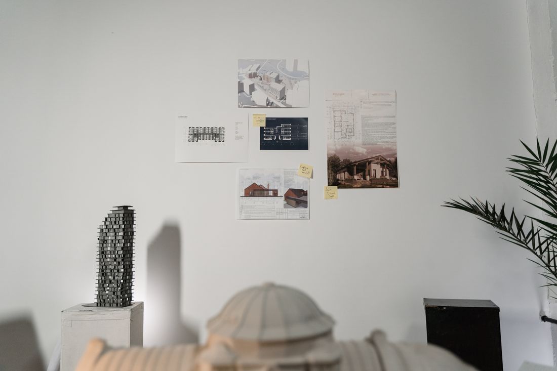 A studio wall pinned with architectural drawings and sketches beside a scale model, with a plant in the corner of a bright design office.