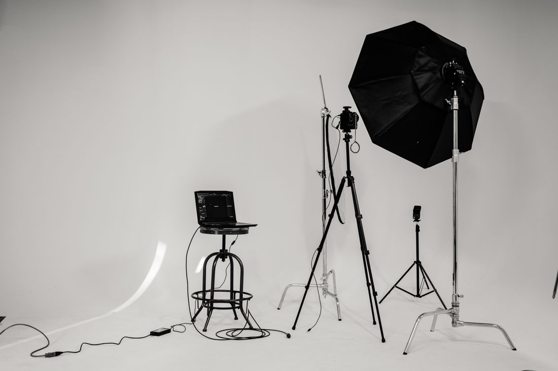 An empty portrait studio corner with a softbox light, a camera on a tripod, a second light stand, and a laptop on a stool against a plain backdrop, no subject in frame.