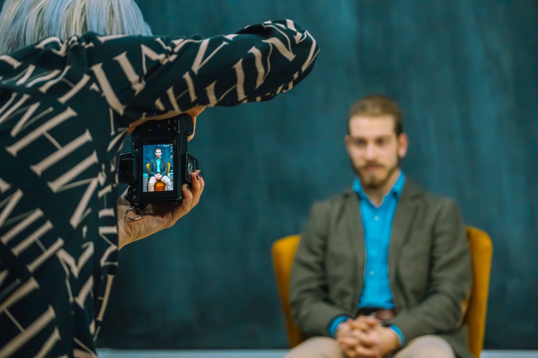A photographer holding a camera up to photograph a man in a blazer and blue shirt, seated with hands clasped against a plain dark teal backdrop, the camera's screen showing the shot in progress.
