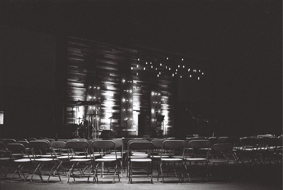 Rows of empty folding chairs face a dimly lit stage with warm accent lighting and hanging bulb strings, house lights low, no audience yet.