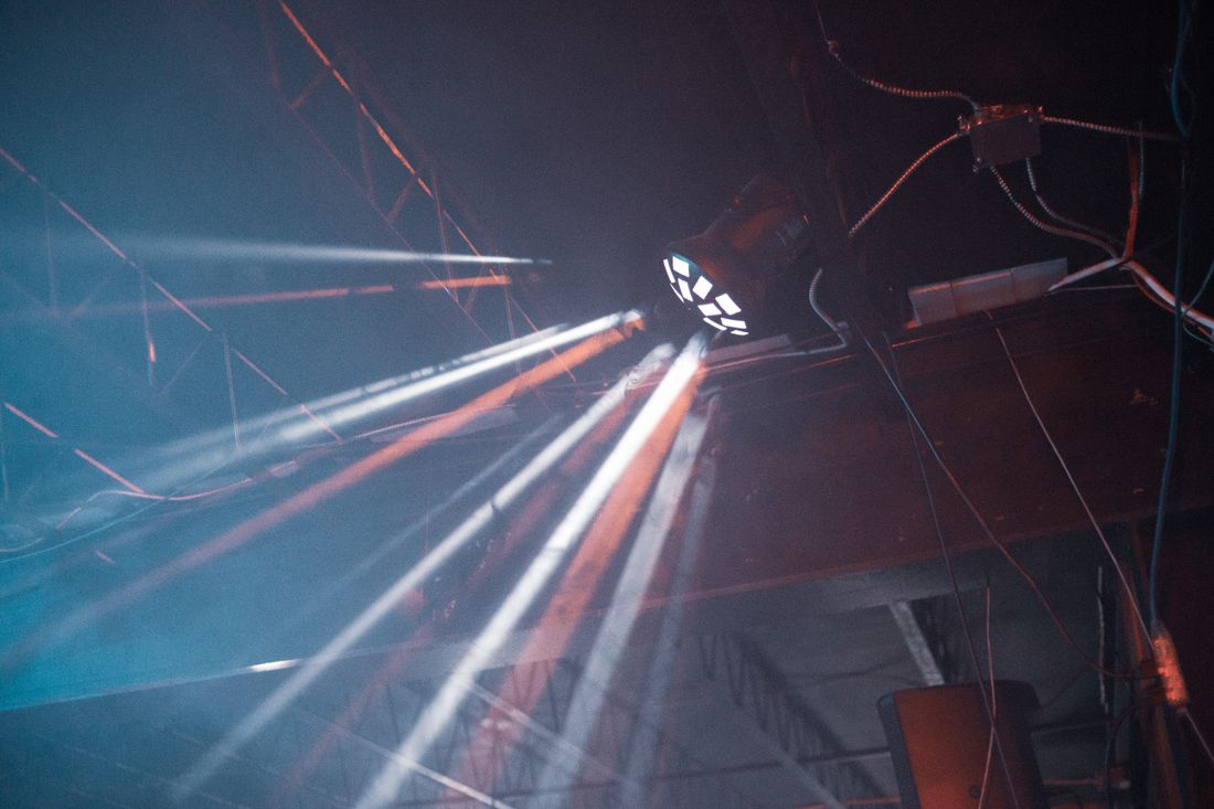 A stage light fixture mounted on an overhead truss casts beams downward against a dark ceiling, seen from below.