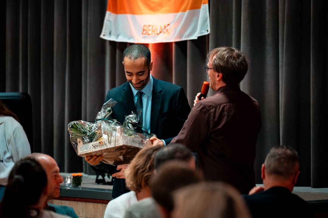 A presenter hands an award to a recipient on stage in front of a seated audience under warm stage lighting.