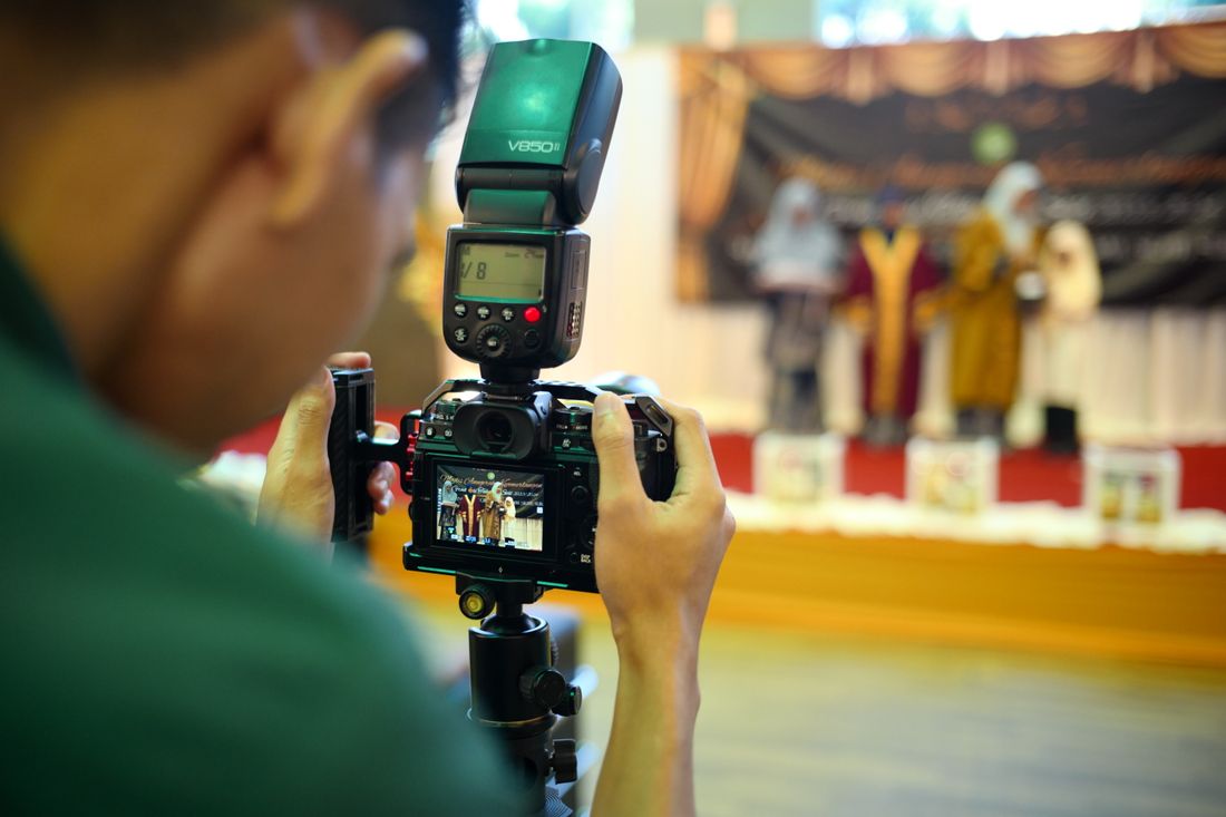 A photographer holding a camera with an on-camera flash, shooting toward a lit stage from the audience side.