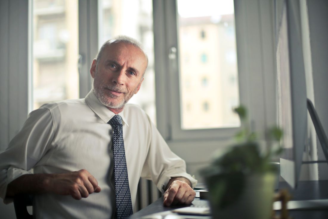 A calm, direct-expression executive portrait of a man in a white collared shirt and tie seated near an office window, mid-distance framing with a soft blurred building view behind him.