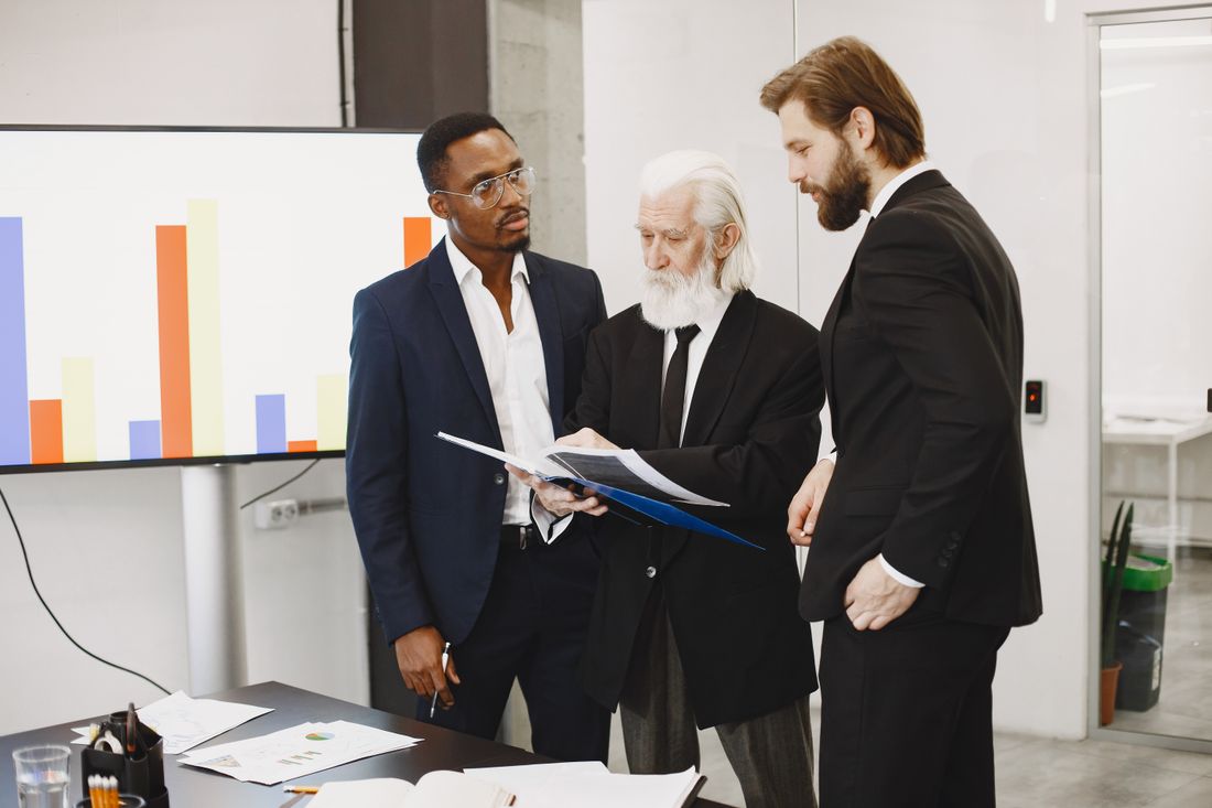 Three professionals in business suits standing together in an office, calmly reviewing a document folder, with a plain bar chart on a wall screen behind them.