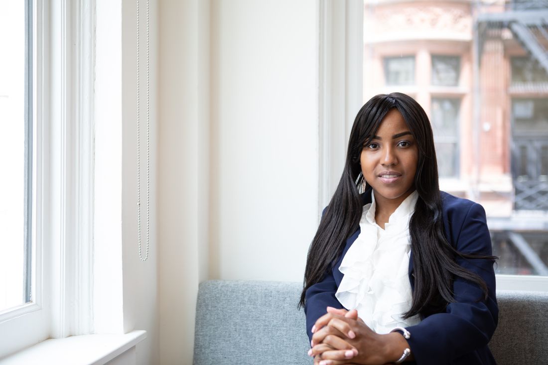 A businesswoman in a navy blazer sits calmly on a sofa near a large window in a bright office lobby, hands clasped, looking toward the camera.