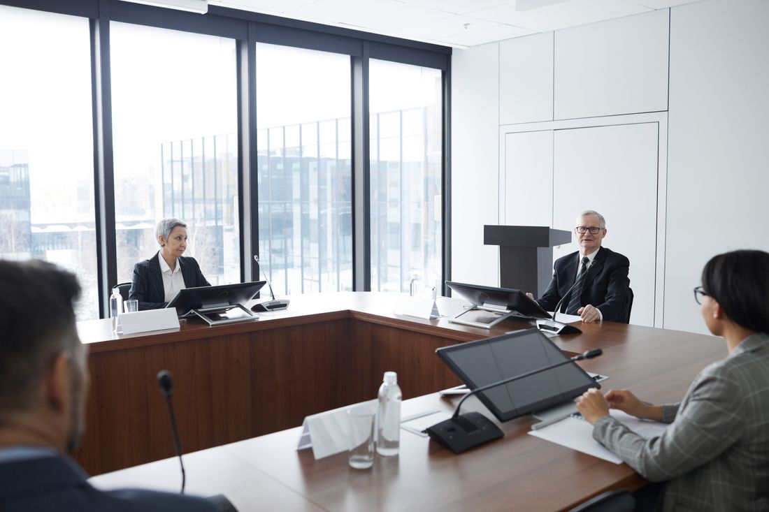 Executives in business attire seated around a formal boardroom table near large windows during a meeting.