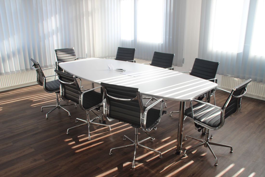 A small private meeting room with a polished white table and black high-back chairs arranged closely together, sunlight streaming through sheer window coverings onto a dark wood floor.