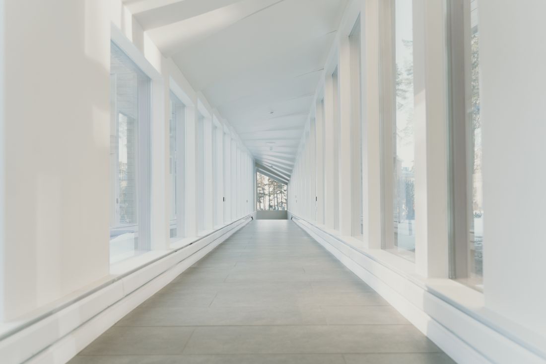 A quiet, empty covered corridor lined with windows on both sides, soft daylight coming in from a window at the far end.