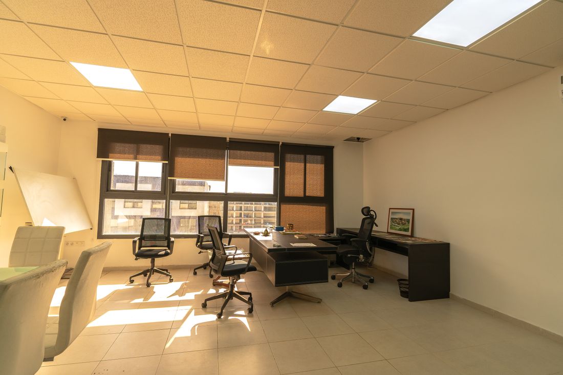 A small, quiet office with desks, chairs, and daylight streaming in through a bank of windows.