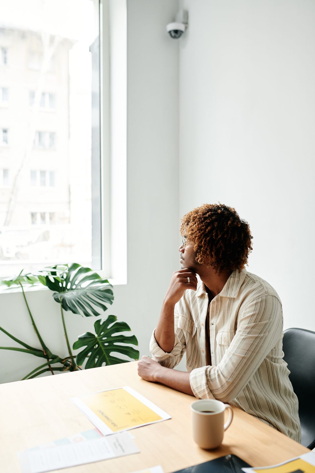 A professional sits at a desk with a coffee mug and blurred paperwork, pausing to look out the window in thought.