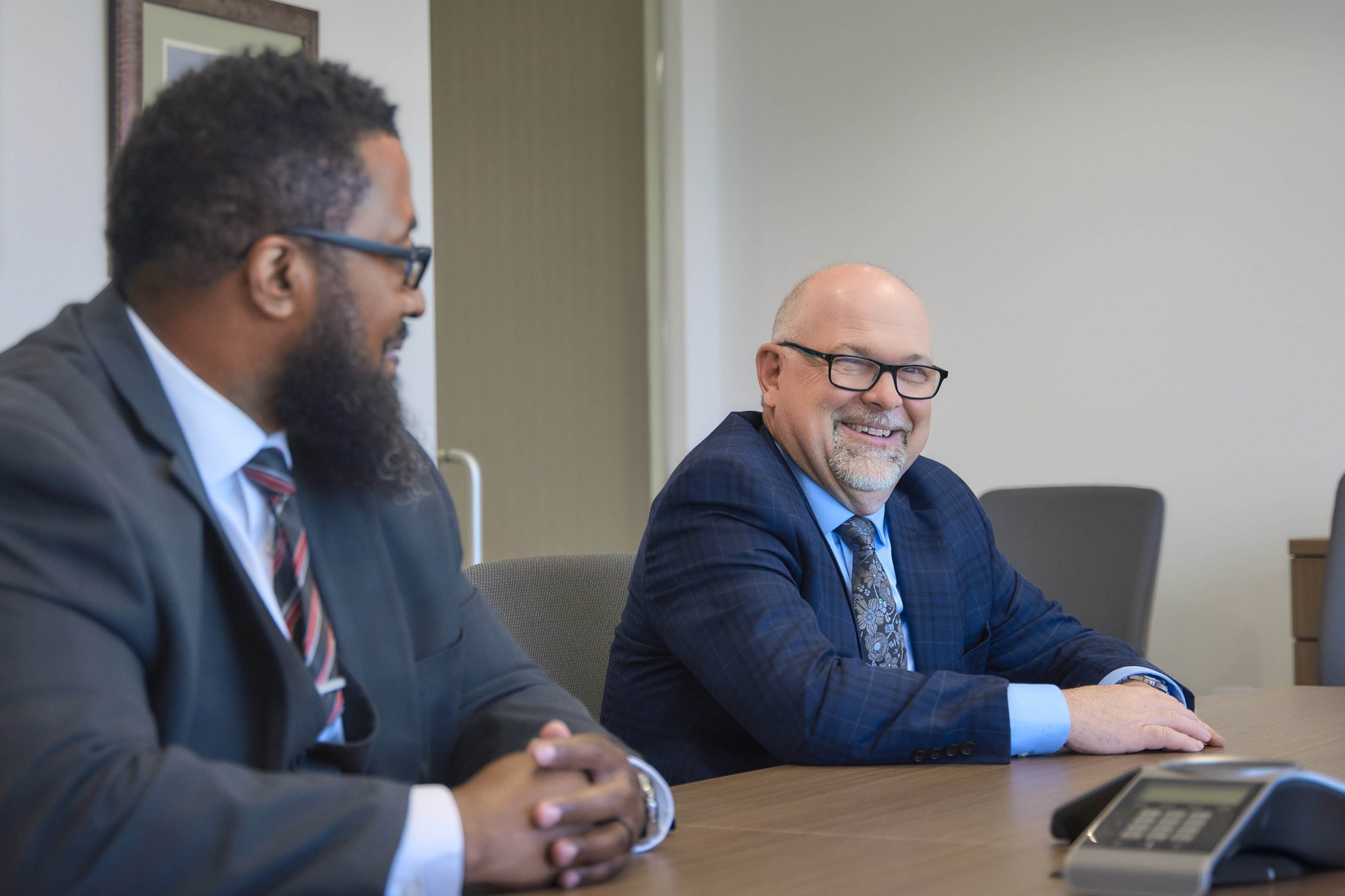 Two men in business suits sit at a conference table talking, one smiling and looking toward the camera.