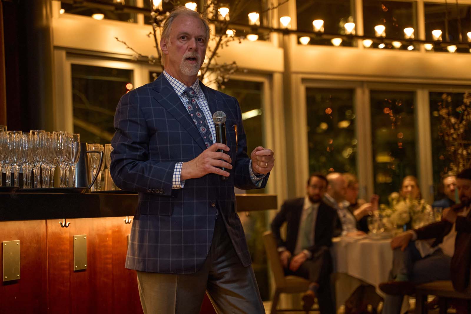 Man in a plaid blazer speaks into a microphone at an indoor event with string lights and seated guests behind him, the kind of moment a brief should flag as must-not-miss