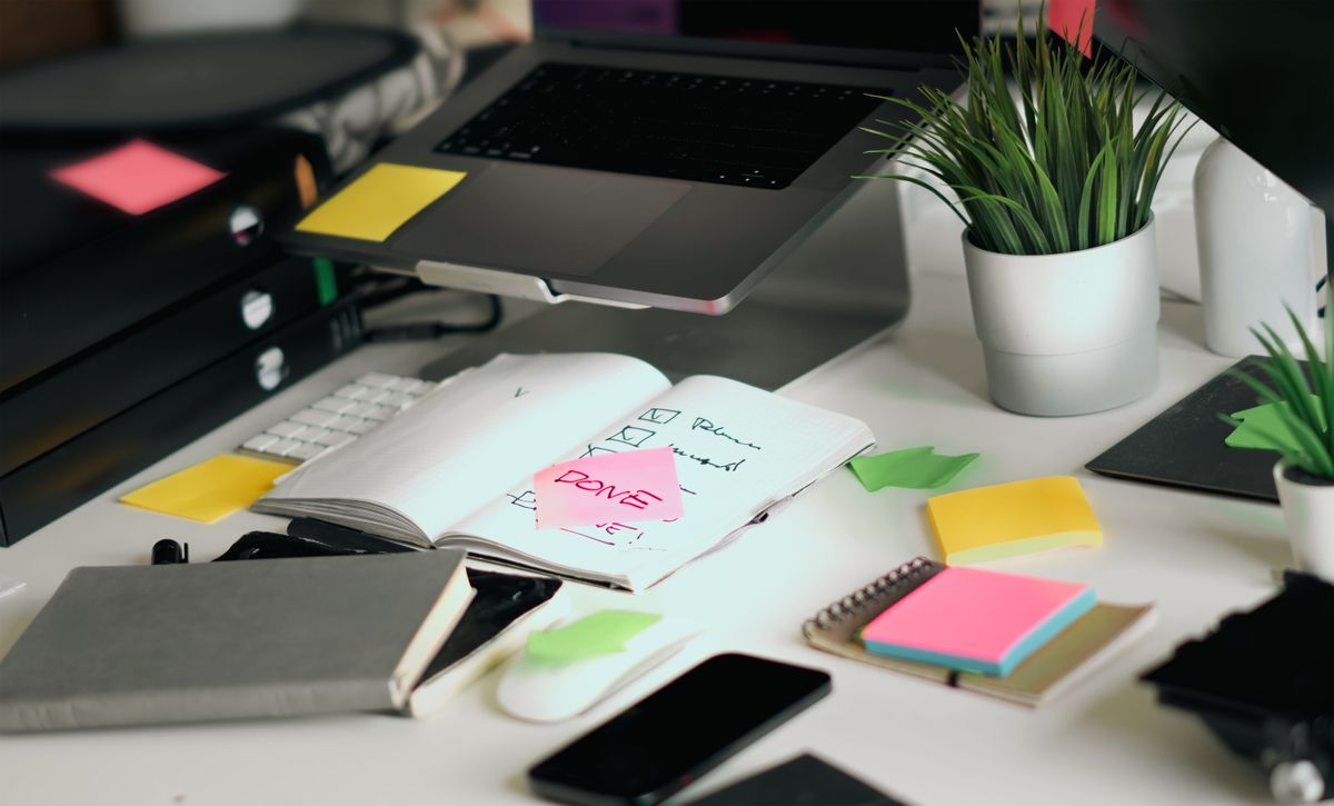 A cluttered desk with a laptop, notebook, checklist, and colorful sticky notes, representing planning notes for an event photography brief