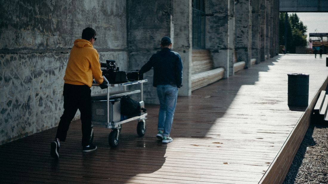 Two crew members pushing an equipment cart loaded with camera gear cases along an outdoor walkway, seen from behind.