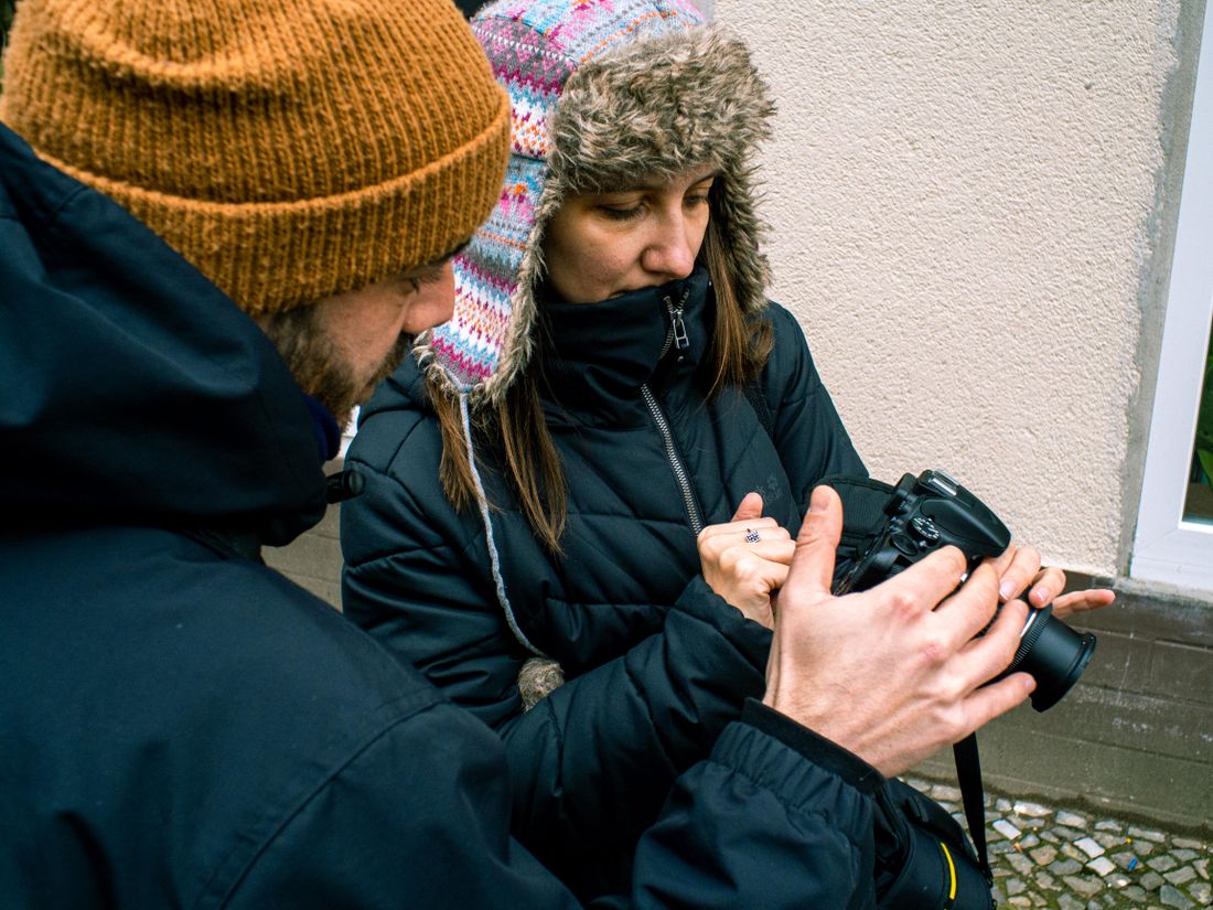 Two photographers in dark jackets standing outdoors near a plain building wall, one turning a camera's rear screen toward the other to show a shot.
