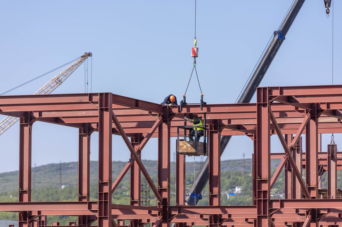 A construction worker in a hard hat rides a crane-lifted platform beside exposed red structural steel framing on a high-rise job site, with a second crane visible against a clear sky.
