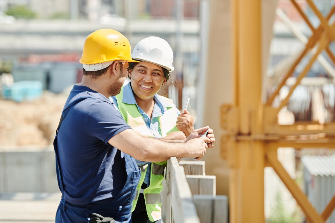 Two people in hard hats, one wearing a safety vest and holding a small tool, talk near a wooden railing at an elevated construction site with a tower crane and city skyline in the background.