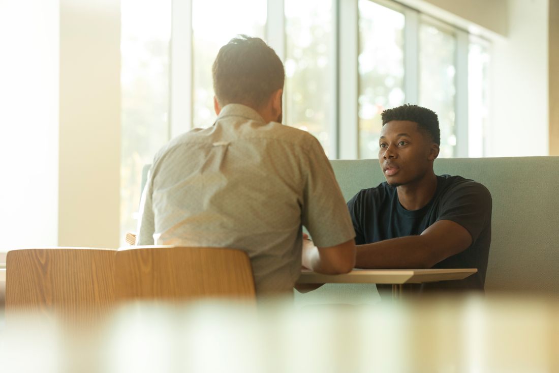 An older adult seen from behind talking with a younger adult across a small table beside tall windows in a bright interior.