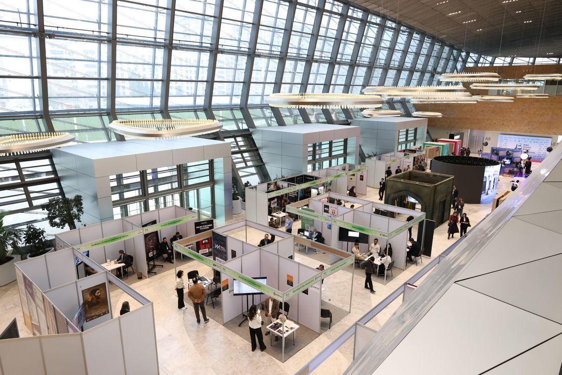 A wide overhead view of an exhibition hall with rows of small open booths, tables, and people talking, under a glass roof.