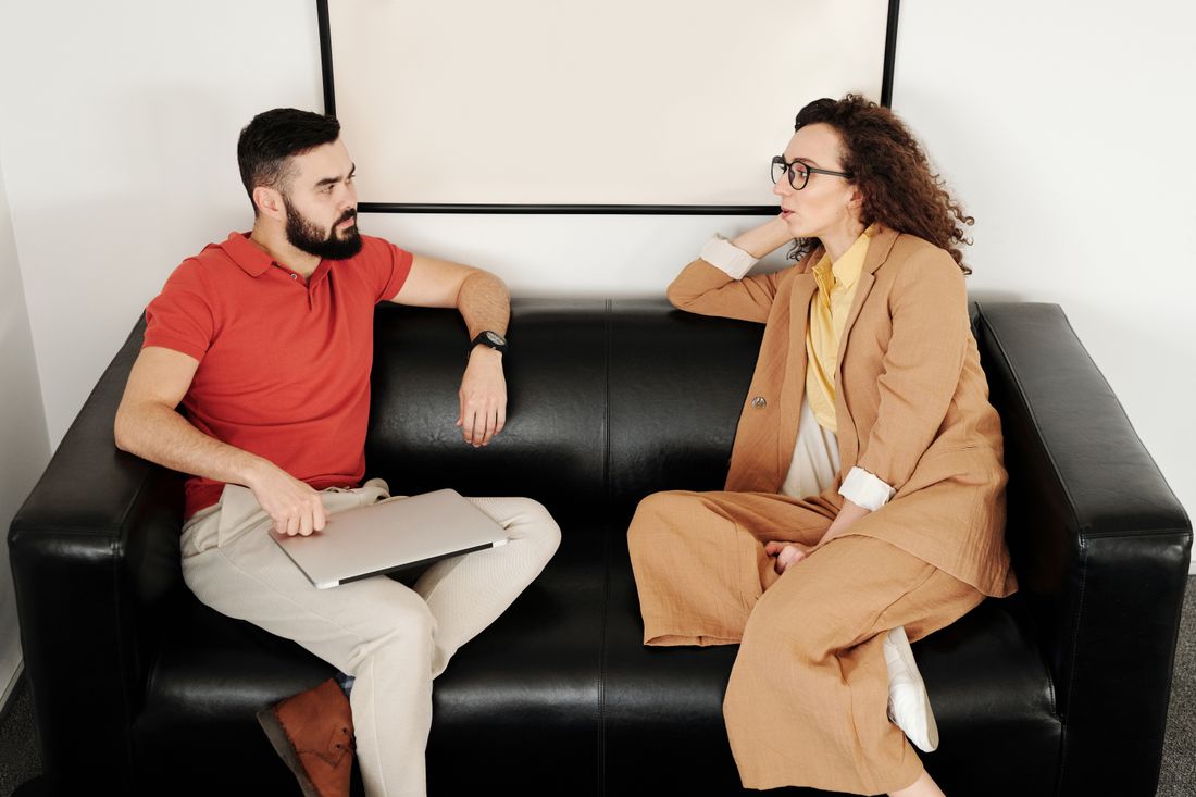 A man and woman having a conversation on a black sofa in a plain room.