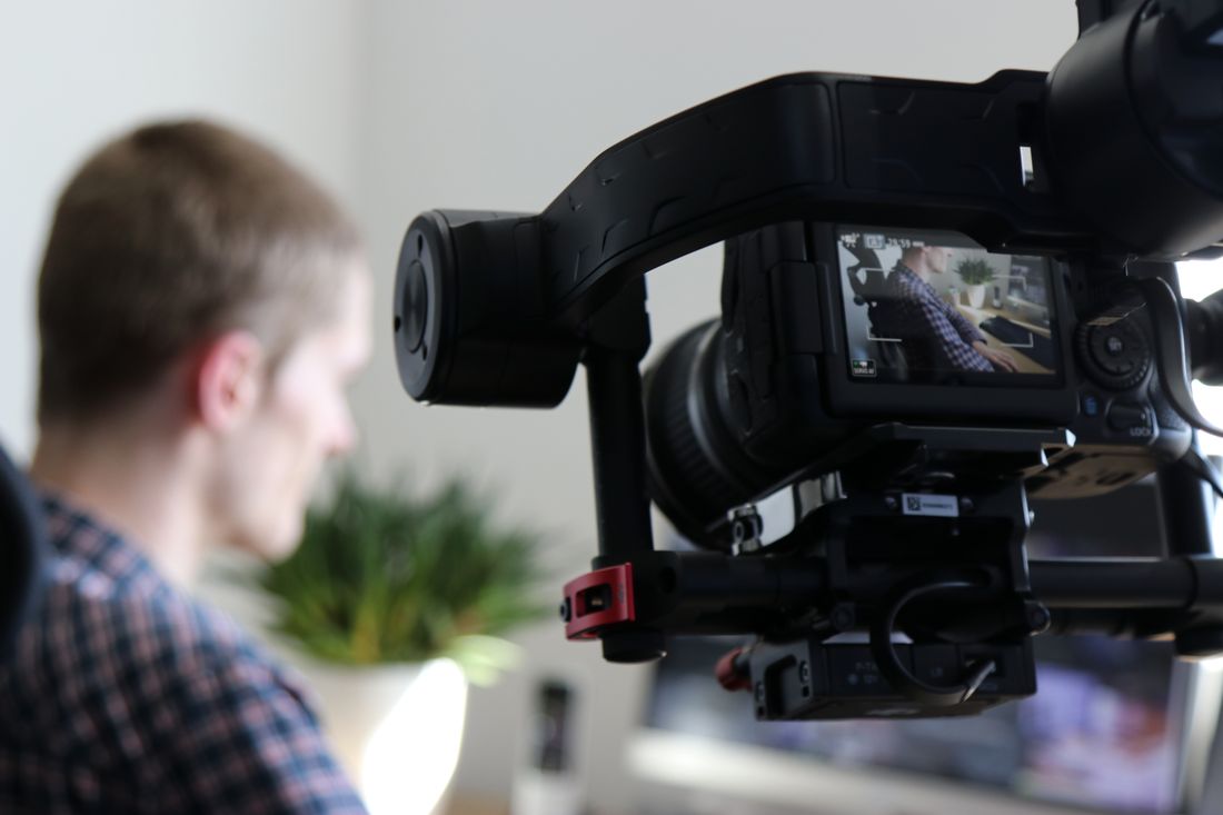 A video camera in the foreground films a person being interviewed indoors, with the shot visible on the camera's monitor.