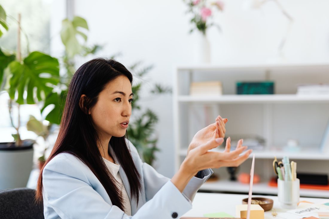 A woman in a light blazer gestures with both hands while talking, seated at a desk in a bright office with plants and a bookshelf in the background.