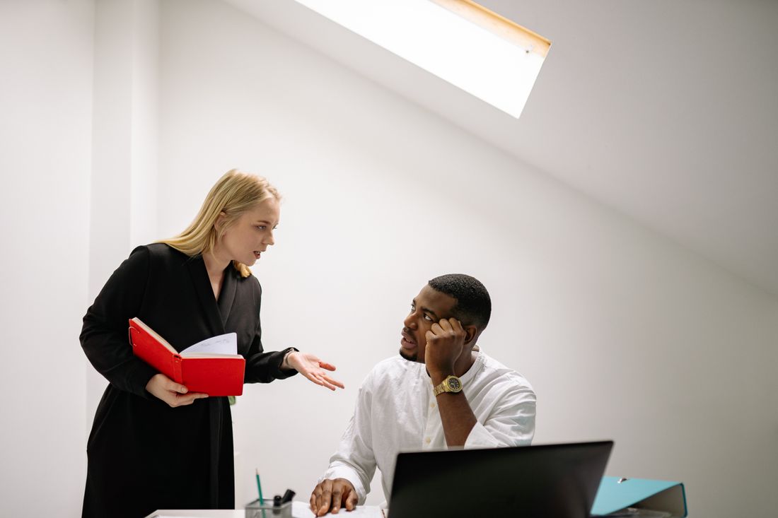 A woman holding an open notebook gestures with one hand while explaining something to a man seated at a desk with a laptop, in a plain white room with a skylight.