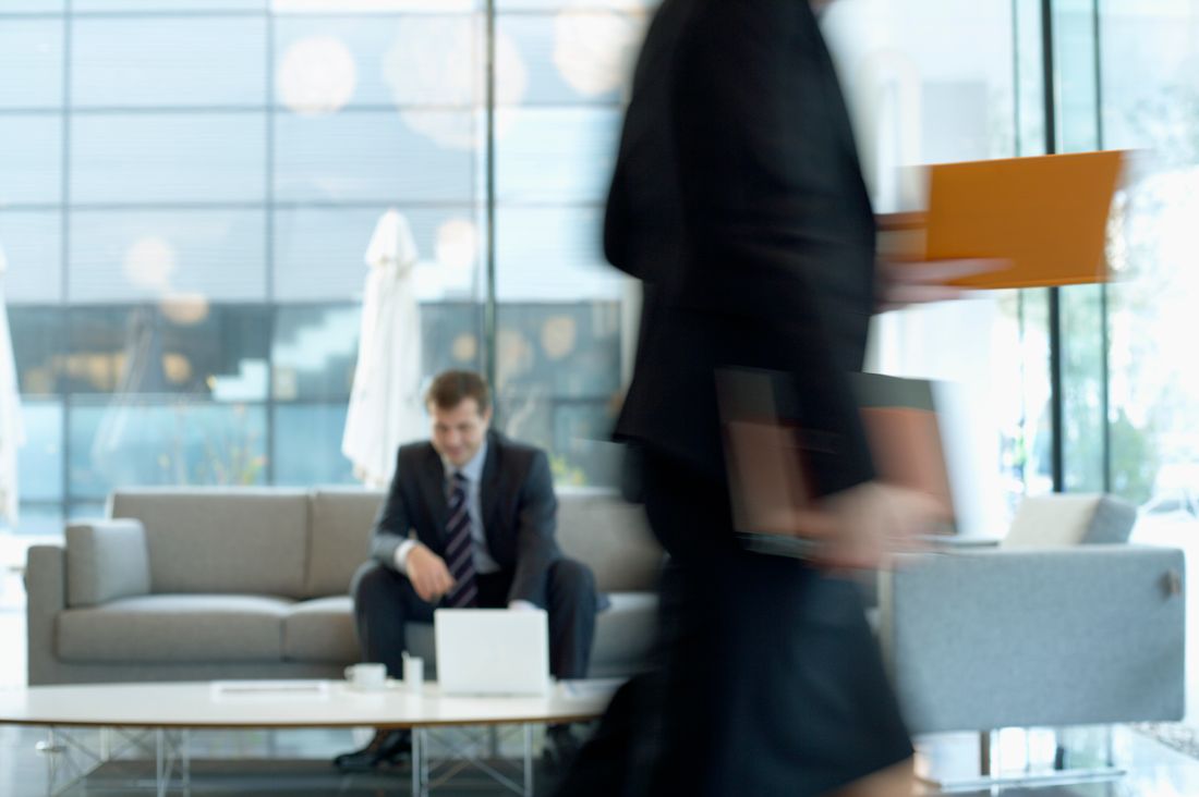 A man in a suit sits in focus on a lounge sofa in a glass-walled office while a colleague carrying folders and a laptop blurs past in the foreground.