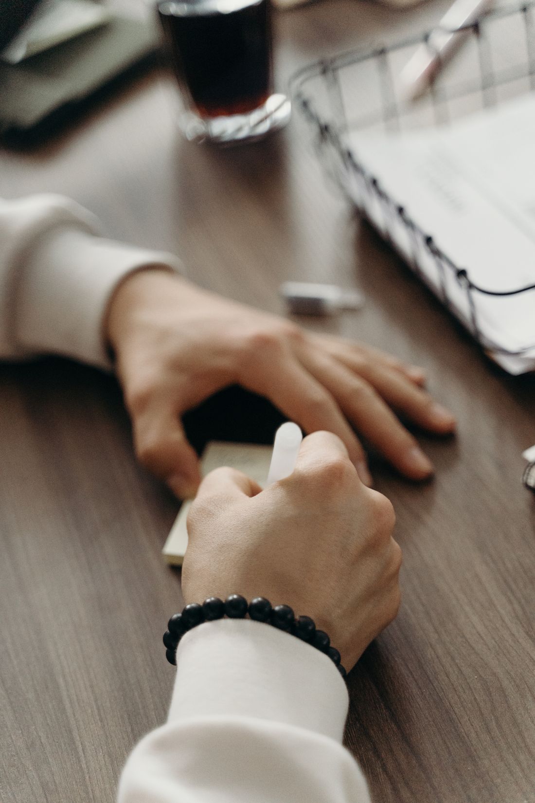 Close-up of a hand writing in a small notebook at a wooden desk, with a coffee cup and a wire document tray nearby.